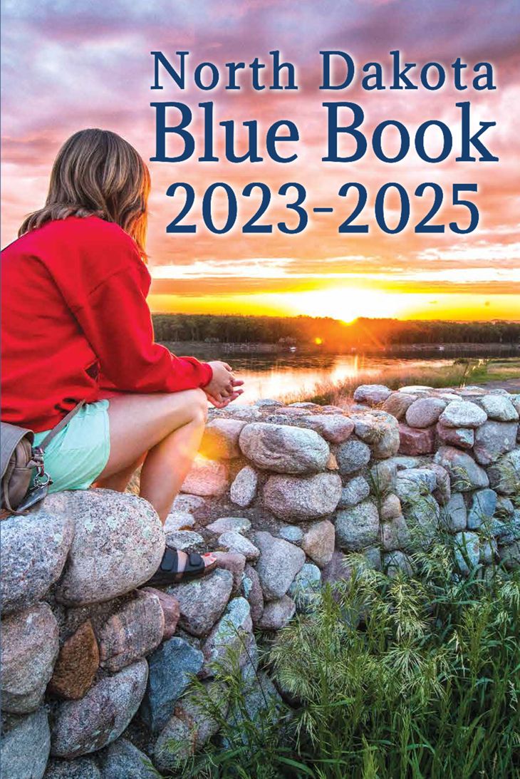 North Dakota Blue Book Cover - girl sitting on rock wall overlooking a sunset on the Missouri River