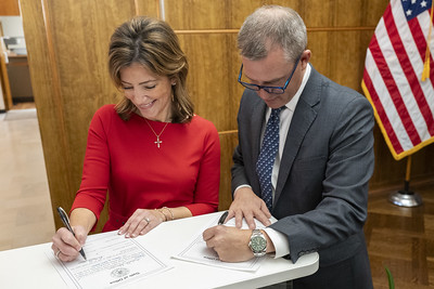 Lieutenant governor Strinden is standing on the left side of the photo while signing her oath of office. She is a white middle-aged female with shoulder length brown hair and is wearing a red dress with sleeves and a cross necklace. Governor Armstrong is standing on the right side of Strinden and is signing his oath of office. He is a white middle-aged male with gray hair and is wearing a gray suit and blue tie. 